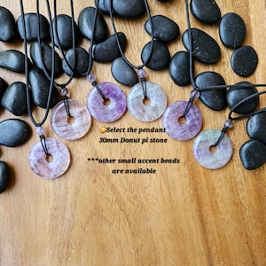 Könnte beinhalten: Fünf Halsketten mit donutförmigen Amethyst-Anhängern an schwarzen Schnüren. Die Anhänger haben einen Durchmesser von etwa 30 mm und ein Loch in der Mitte. Der Text "Select the pendant 30mm Donut pi stone ***other small accent beads are available" ist im Bild sichtbar.