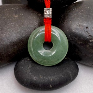 May include: A green jade donut pendant with a hole in the center, hanging from a red cord with a silver bead. The pendant is resting on a pile of black stones.