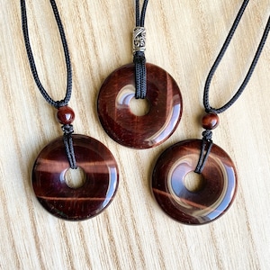 May include: Three circular pendants made of polished dark red tiger eye stone, each featuring a central hole. The pendants are suspended on black cord necklaces, with two featuring small matching stone beads and one featuring a silver-toned metal bead.
