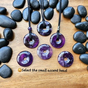 May include: Six purple amethyst donut pendants on black cords, with a small silver accent bead on one of the cords. The pendants are arranged on a wooden surface with black river rocks.