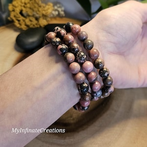 May include: Three stacked beaded bracelets featuring round, polished stones in shades of dusty pink, mottled grey, and black. The beads are strung on an elastic cord. The text MyInfinateCreations is visible in the lower left corner of the image.
