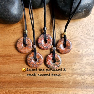 Puede incluir: Cinco collares con colgantes redondos con patrones rojos, naranjas y marrones y pequeñas cuentas de acento doradas y plateadas. Los collares están sobre una superficie de madera con dos piedras negras.