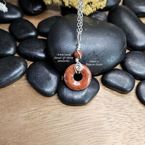 Op de afbeelding: Een hanger met een 18 mm donut pi-steen in rood-bruine tinten, hangend aan een zilveren ketting. De hanger heeft een cirkelvormig ontwerp met een centraal gat en wordt geaccentueerd door een kleinere, ronde steen. De tekst luidt "Select a Rope or Chain".