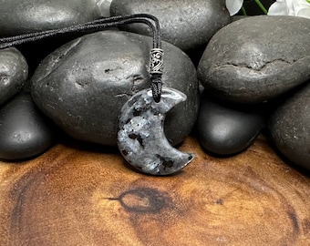 May include: A crescent moon-shaped pendant made of speckled gray and black stone with iridescent flecks. The pendant hangs from a black cord featuring a decorative silver-toned metal bead with an intricate scrollwork pattern at the top.