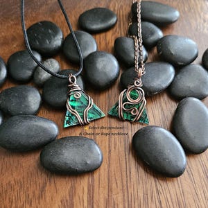 May include: Two triangular green stone pendants with copper wire wrapping, one on a black rope necklace and the other on a copper chain. The pendants are displayed on a wooden surface with black stones. Text reads: "Select the pendant & Chain or Rope necklace."