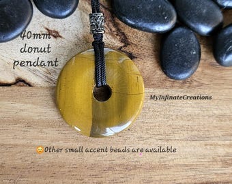 May include: A 40mm donut pendant made of tiger's eye stone, with a black cord. The pendant is a golden brown color with dark brown stripes. The text "MyInfiniteCreations" is visible on the right side.