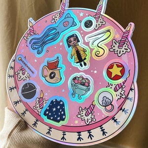 Puede incluir: Un pastel hologr&aacute;fico rosa con varias pegatinas en &eacute;l. Las pegatinas incluyen un calamar, una ni&ntilde;a con un impermeable amarillo, un rat&oacute;n con un sombrero, un taz&oacute;n de caramelos, una estrella, una concha, un su&eacute;ter y una varita.