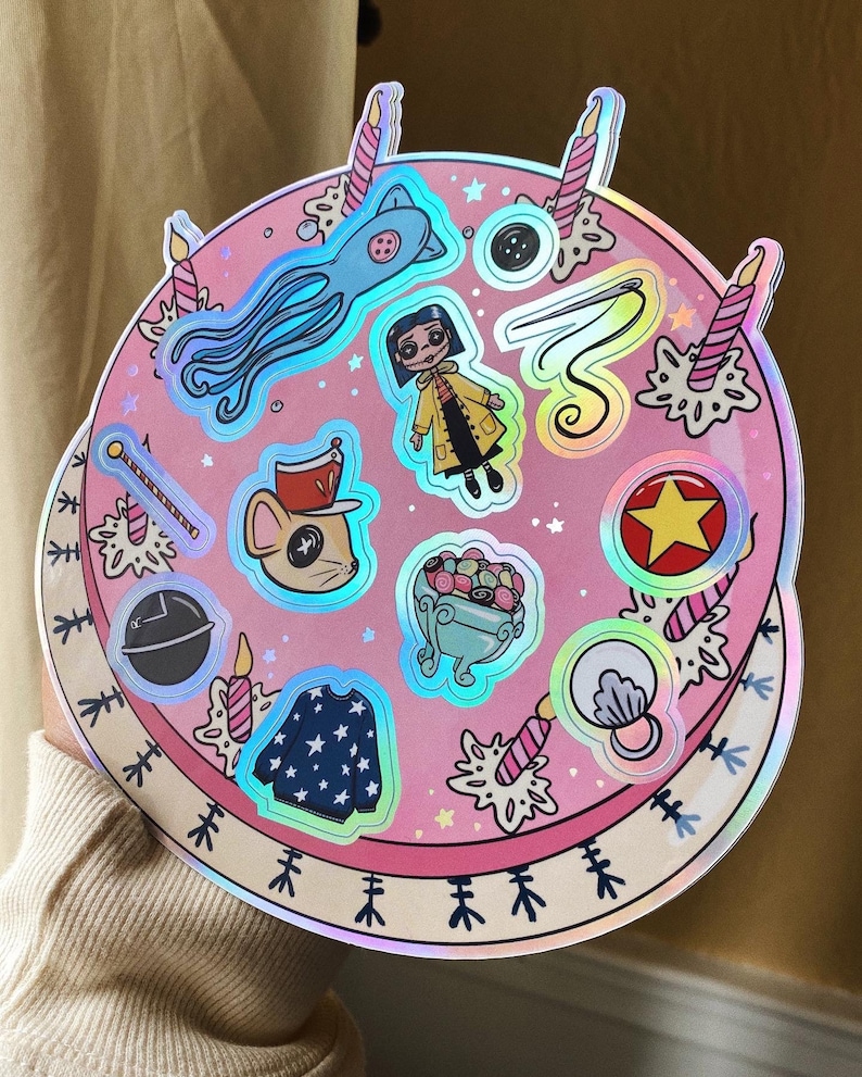 Puede incluir: Un pastel hologr&aacute;fico rosa con varias pegatinas en &eacute;l. Las pegatinas incluyen un calamar, una ni&ntilde;a con un impermeable amarillo, un rat&oacute;n con un sombrero, un taz&oacute;n de caramelos, una estrella, una concha, un su&eacute;ter y una varita.