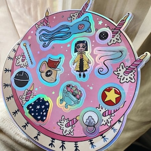 Puede incluir: Una hoja de pegatinas circular rosa y blanca con acentos hologr&aacute;ficos. La hoja presenta varios personajes y objetos de la pel&iacute;cula "Coraline", incluyendo a Coraline, la Otra Madre, el gato y un rat&oacute;n con ojos de bot&oacute;n.