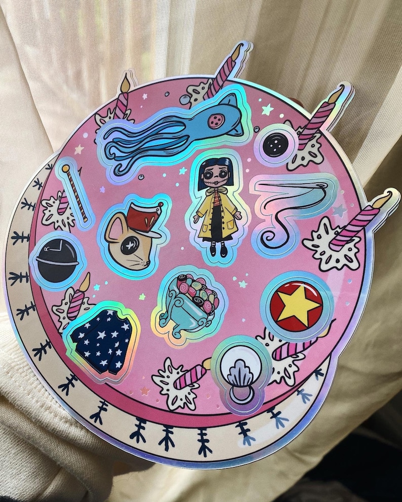 Puede incluir: Una hoja de pegatinas circular rosa y blanca con acentos hologr&aacute;ficos. La hoja presenta varios personajes y objetos de la pel&iacute;cula "Coraline", incluyendo a Coraline, la Otra Madre, el gato y un rat&oacute;n con ojos de bot&oacute;n.