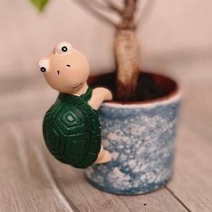 Puede incluir: Una pequeña figura de tortuga verde con cabeza marrón y ojos blancos está trepando por el lado de una maceta de cerámica azul y blanca. La maceta tiene un borde marrón y está llena de tierra marrón oscura.