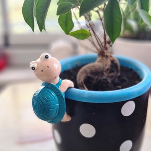 May include: A small, blue turtle figurine sits on the edge of a black and white polka dot planter. The planter is filled with soil and a small green plant with brown roots.