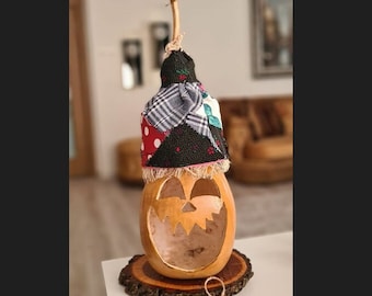 Può includere: Una decorazione di Halloween fatta a mano realizzata con una zucca con un viso di zucca intagliato. La zucca è sormontata da un cappello di stoffa colorata a patchwork e poggia su una base di legno. La base ha un piccolo cartellino con la scritta "fatto a mano con amore".