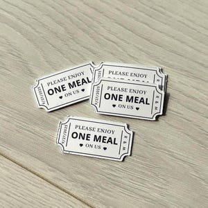 May include: A collection of white and black meal tickets with a rounded design, resting on a light wood surface. Each ticket displays the text "PLEASE ENJOY ONE MEAL ON US" with a heart symbol. The date "9/27/2025" is printed on each ticket.