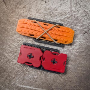 Peut inclure: Une plaque de désensablement orange vif à surface texturée fixée par un cordon élastique beige, placée au-dessus de deux petits bidons de carburant rouges montés sur une base rectangulaire noire avec des détails moulés et des boutons noirs.