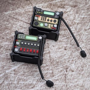 Puede incluir: Dos dispositivos de comunicación por radio en miniatura de color negro alojados en marcos protectores. Cada unidad cuenta con una pantalla verde, un teclado numérico, varios botones circulares y un cable negro en espiral.