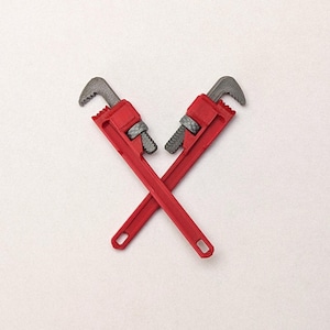 May include: Two red pipe wrenches with grey metal jaws and adjustment nuts, positioned in a crossed formation. Each tool features a long handle with a small rectangular hole at the end, showcasing a distinct contrast between the red and grey.