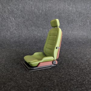Peut inclure: Un siège de voiture miniature dans les tons de vert, marron et noir. Le siège est doté d'un appui-tête et d'un design profilé. Le dos du siège est marron et la base est noire. Le siège est conçu pour ressembler à un siège de voiture vintage.