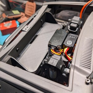 Peut inclure: Gros plan sur le compartiment moteur gris d'une voiture miniature. Le compartiment contient des composants électroniques noirs, des câbles et un panneau gris texturé. Le compartiment moteur est ouvert, révélant la mécanique interne du modèle.