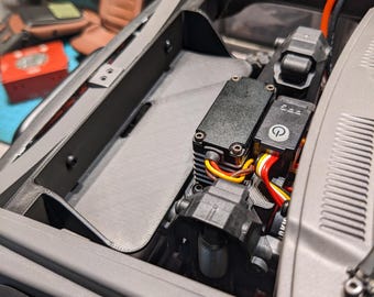 May include: Close-up of a gray model car engine compartment. The compartment contains black electronic components, wiring, and a gray textured panel. The engine bay is open, revealing the internal mechanics of the model.