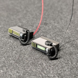 May include: Two miniature grey walkie-talkies with black coiled cords and aerials. Each radio has a small screen and a black microphone. One radio has a red wire, and the other has a black wire. The radios are on a grey surface.