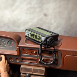 Peut inclure: Une radio miniature noire et verte avec le chiffre 25 affiché à l'écran. La radio est connectée à un microphone noir avec un cordon enroulé. La radio est placée sur un tableau de bord marron.