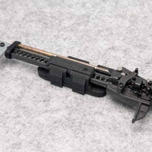 May include: A mechanical assembly featuring a black textured plastic housing, a central metallic rod with a copper-toned finish, and a series of small silver-colored screws. The device includes a notched rail system and various articulated components.