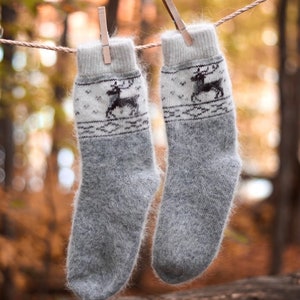 Puede incluir: Dos calcetines de lana gris con un diseño de reno colgados de un tendedero con pinzas de madera. Los calcetines son esponjosos y tienen una banda blanca con un diseño de reno negro. El fondo es un bosque borroso.