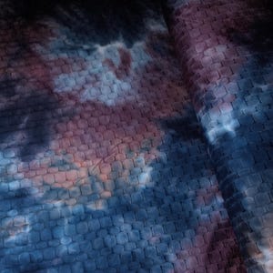May include: Close-up of a textured fabric with a pattern of small, square tiles. The fabric features a blend of colors, including deep blues, purples, and hints of orange, creating a tie-dye effect. The material appears to be folded.