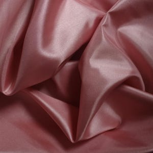 May include: Close-up of a folded piece of pink satin fabric. The fabric has a smooth, shiny surface with visible folds and shadows, creating a sense of depth and texture. The color is a soft, muted pink.