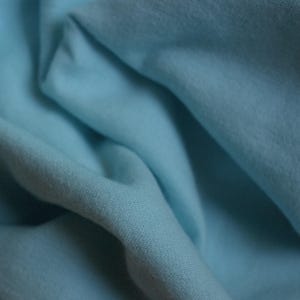 May include: Close-up of a light blue fabric with soft folds and a textured surface. The fabric appears to be a lightweight material, possibly cotton or linen, with a subtle sheen.