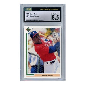 Puede incluir: Una tarjeta de béisbol Upper Deck de 1991 con Michael Jordan en un uniforme de béisbol rojo y blanco, balanceando un bate. La tarjeta está calificada con un 8.5 por CSG.