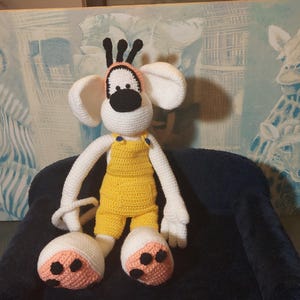 Könnte beinhalten: Handgemachte Häkelpuppe mit weißem Kopf, großen Ohren, schwarzer Nase und gelbem Overall. Die Puppe hat weiße Arme und Beine sowie große Füße mit rosa und schwarzen Akzenten. Sie sitzt auf einer dunkelblauen Oberfläche.