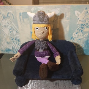 Könnte beinhalten: Handgefertigte Häkelpuppe einer Wikingerfigur, die einen grauen Helm mit weißen Hörnern, ein lila Oberteil und braune Stiefel trägt. Die Puppe hat gelbes Haar und sitzt auf einem kleinen, dunkelblauen Sofa. Der Hintergrund zeigt ein Gemälde.