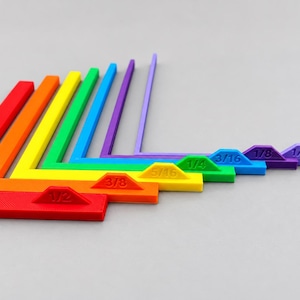 May include: A set of seven L-shaped corner alignment tools in a rainbow color gradient. Each tool features a raised tab with embossed imperial measurements: 1/2, 3/8, 5/16, 1/4, 3/16, 1/8, and 1/16 inches, arranged in descending size order.