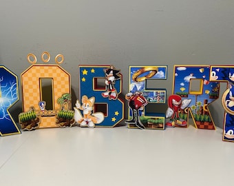 May include: A set of colorful cardboard letters spelling out the name "ROBERT" decorated with characters from the Sonic the Hedgehog video game franchise. The letters are blue, yellow, and red with images of Sonic, Tails, Knuckles, and Shadow.
