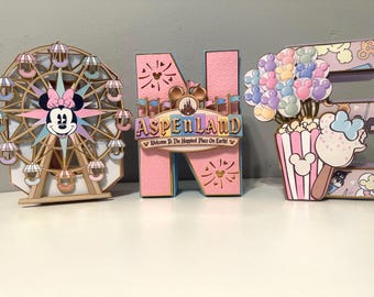 May include: Three decorative letters spelling ONE, featuring a Ferris wheel with a Minnie Mouse face, a pink letter N with a sign reading ASPENLAND Welcome To The Happiest Place On Earth, and a letter E decorated with popcorn and balloons.