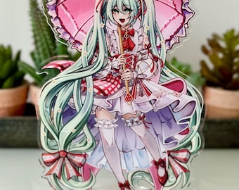 Könnte beinhalten: Ein Anime-Charakter mit grünen Haaren, der ein rosa-weißes Kleid trägt und einen rosa Schirm hält. Der Charakter trägt rote Schuhe und hat eine rote Schleife im Haar. Der Charakter steht auf einem klaren Acrylsockel.