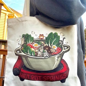 May include: A cream-colored tote bag featuring a colorful illustration of a hot pot with cartoon cats. The hot pot is on a red burner with the words "HOT POT SPRINGS" in white. The bag has a simple design with a rectangular shape and long straps.