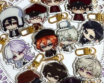 Może przedstawiać: Kolekcja dwunastu przezroczystych akrylowych breloków przedstawiających postacie z anime w stylu chibi o różnych kolorach włosów, strojach i nakryciach głowy. Każdy brelok jest przymocowany do złotego metalowego zapięcia typu karabińczyk.