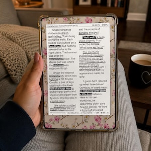 May include: A digital e-reader displaying the text of Percy Jackson Complete Series #1-5, encased in a clear protective cover featuring a delicate pink and white floral pattern. The screen shows highlighted text passages and a battery indicator.