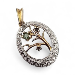 May include: An oval pendant featuring a gold-toned branch motif with a green gemstone and two dark-toned floral accents, set within a silver-toned frame encrusted with numerous small clear stones and a gold-toned bail at the top.