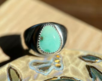 May include: A silver ring with a teardrop-shaped, light green stone. The stone is set in a silver bezel with a scalloped edge. The ring is on a decorative ceramic surface.