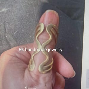 May include: A gold-colored ring with a unique, abstract design. The ring is worn on a finger and is made of metal. The text "Bk handmade jewelry" is visible in the image.
