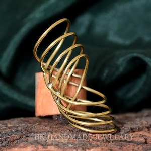 Könnte beinhalten: Ein goldfarbener Metallring mit einem einzigartigen, mehrschichtigen Design. Der Ring besteht aus mehreren ineinander verschlungenen Metallbändern, die einen Spiraleffekt erzeugen. Der Ring wird auf einer Holzoberfläche präsentiert.