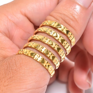 Puede incluir: Anillos dorados con un patrón de corazones apilados en un dedo. Los anillos tienen una superficie texturizada y un diseño de corazón repetido. Los anillos son de color oro cálido y parecen ser de metal.