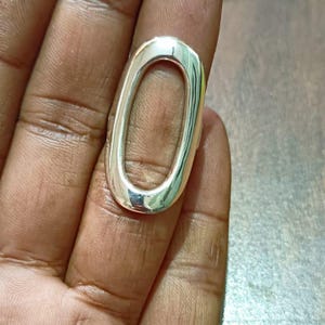Könnte beinhalten: Ein silberner Ring mit einer großen ovalen Öffnung. Der Ring ist dick und hat eine glatte, polierte Oberfläche.