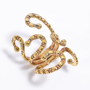 May include: A gold-colored ring with an abstract design. The ring features multiple curved, textured bands that loop and swirl, creating an open, sculptural form. The metal has a slightly aged appearance.