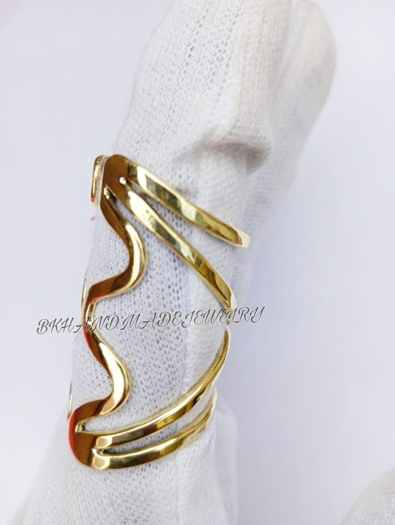 May include: Gold-toned metal ring with a wavy design. The ring is made of multiple bands that are intertwined. The ring is on a white surface. "BKH AND MADE JEWELRY" is written on the surface.