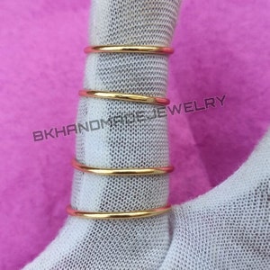 May include: Four gold-colored metal rings stacked on top of each other. The rings are smooth and have a shiny finish. The rings are on a white fabric background.